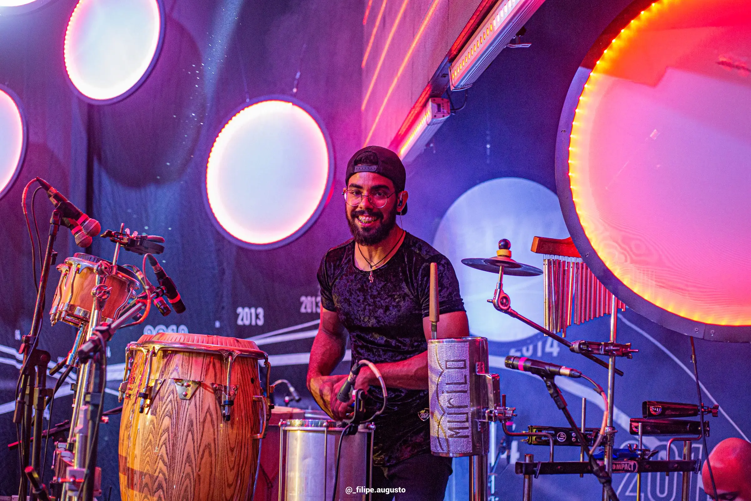 Murilo Pontes tocando conga no palco, sob luz púrpura.